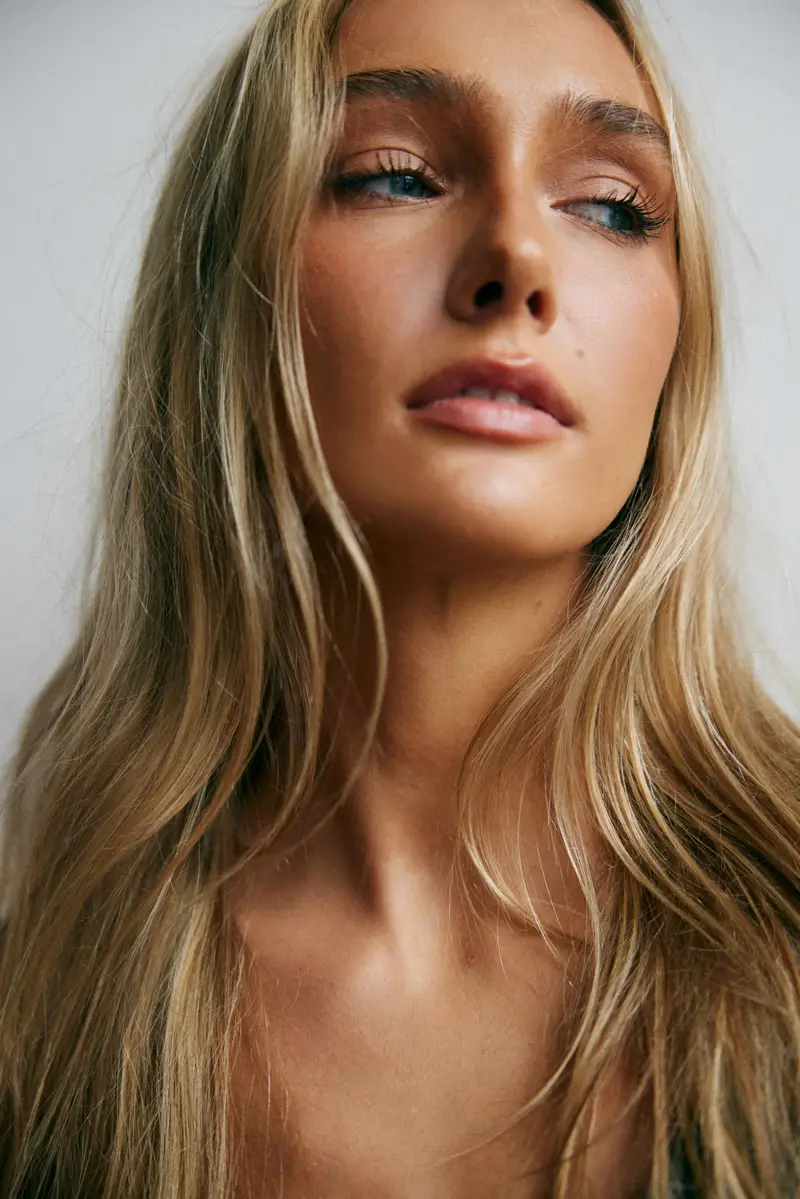 Hannah Caddo in a close-cropped head-and-shoulders portrait, chin lifted and head tilted back, eyes looking past the camera and lips parted, warm bronzed makeup and long wavy blonde hair over bare shoulders, lit by soft daylight against a pale grey wall.