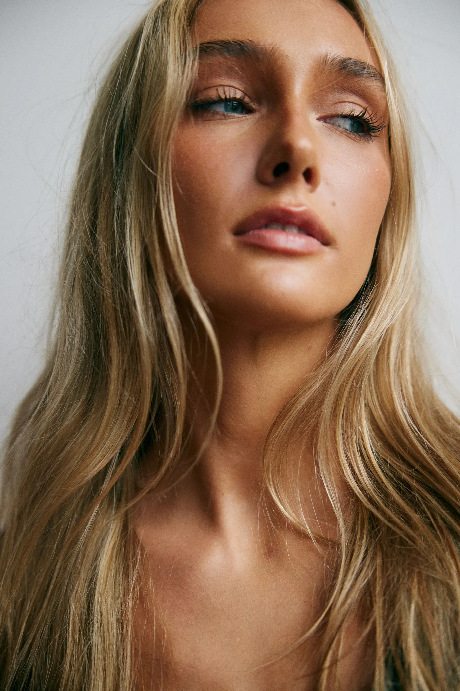 Hannah Caddo in a close-cropped head-and-shoulders portrait, chin lifted and head tilted back, eyes looking past the camera and lips parted, warm bronzed makeup and long wavy blonde hair over bare shoulders, lit by soft daylight against a pale grey wall.