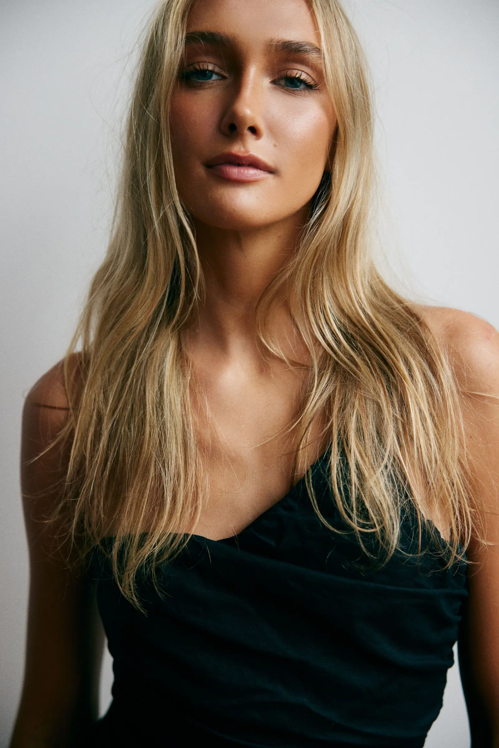 Hannah Caddo framed from the chest up against a grey studio wall in a black asymmetric top, her long pale blonde hair falling loose past her shoulders as she meets the camera with a neutral, closed-lip expression in soft directional light.