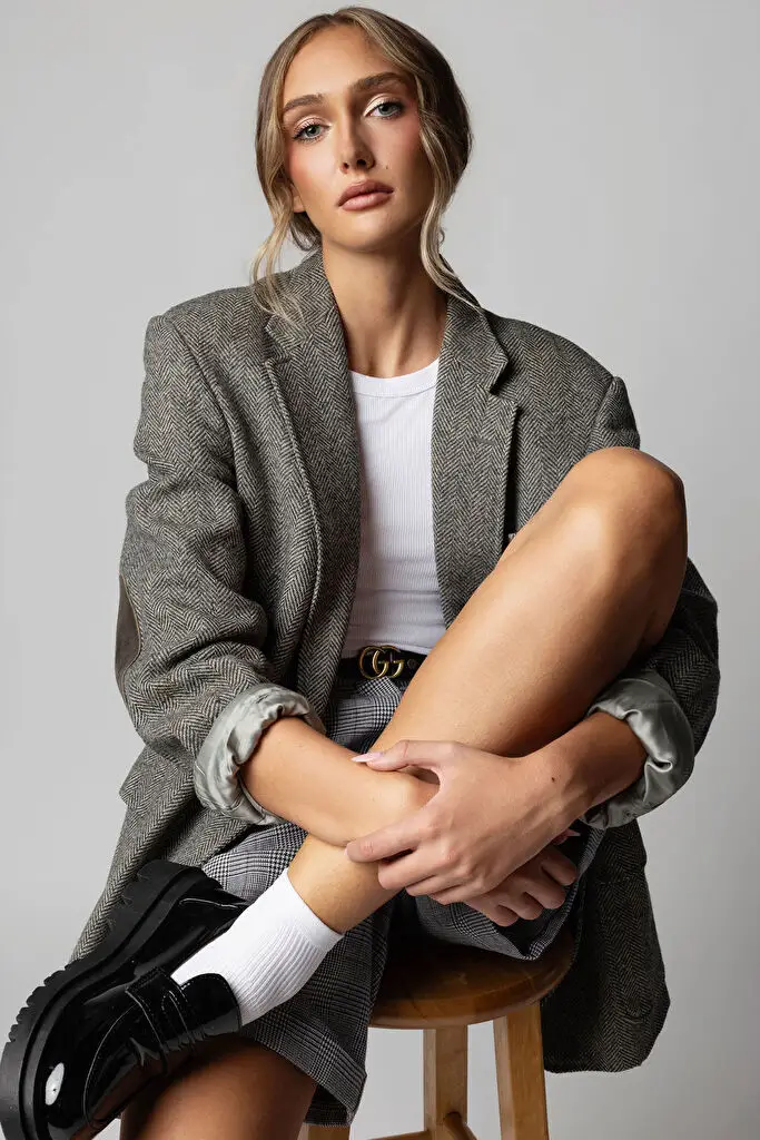Hannah Caddo sits on a stool against a grey backdrop, one leg drawn up, hands clasped at the shin, in an oversized grey herringbone blazer, white tank, plaid mini skirt and black patent loafers, blonde hair swept back, looking at camera in soft light.