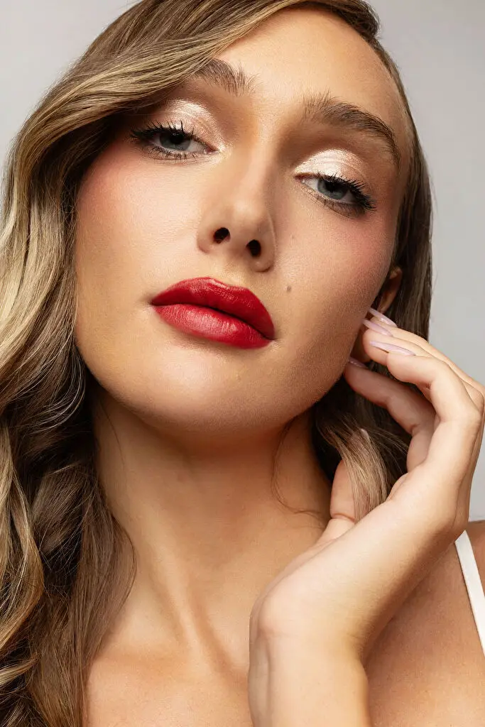 Hannah Caddo in a beauty close-up cropped at the collarbone, chin raised and fingertips resting against her jaw, eyes on the camera; matte red lipstick, pearly champagne eyeshadow and long wavy dark blonde hair, lit evenly by studio light on a grey backdrop.