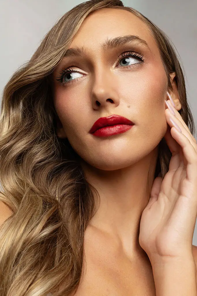 Hannah Caddo, cropped close to head and bare shoulders on a pale grey seamless, tips her chin up with eyes turned away from the lens and fingertips resting at her jaw, wearing glossy red lipstick, long blonde waves falling in soft even light.