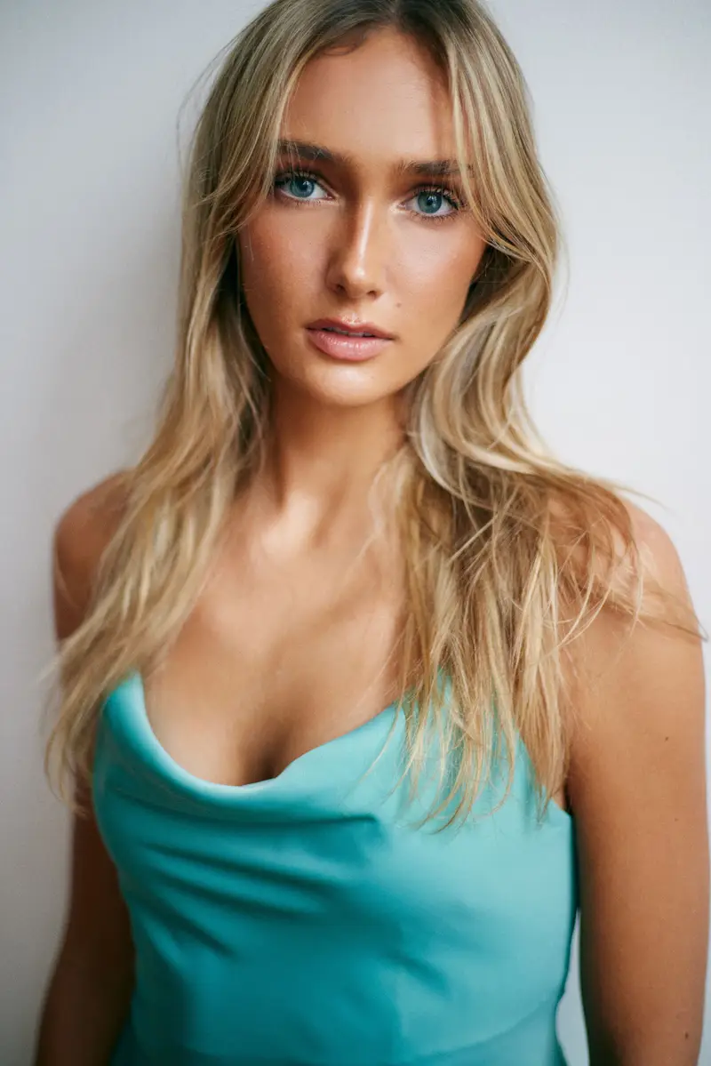 Hannah Caddo faces the camera in a close head-and-chest crop, wearing a draped aqua satin cowl-neck top, long blonde hair falling in loose waves, blue-green eyes level to the lens and freckles visible under dewy makeup, lit by soft daylight against an off-white wall.