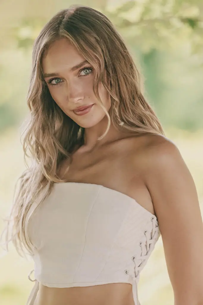 Hannah Caddo photographed from the waist up outdoors, turned toward the camera with a closed-lip smile, wearing a cream bandeau corset top laced through side eyelets, her long wavy dark blonde hair falling loose in warm diffused daylight against blurred foliage.