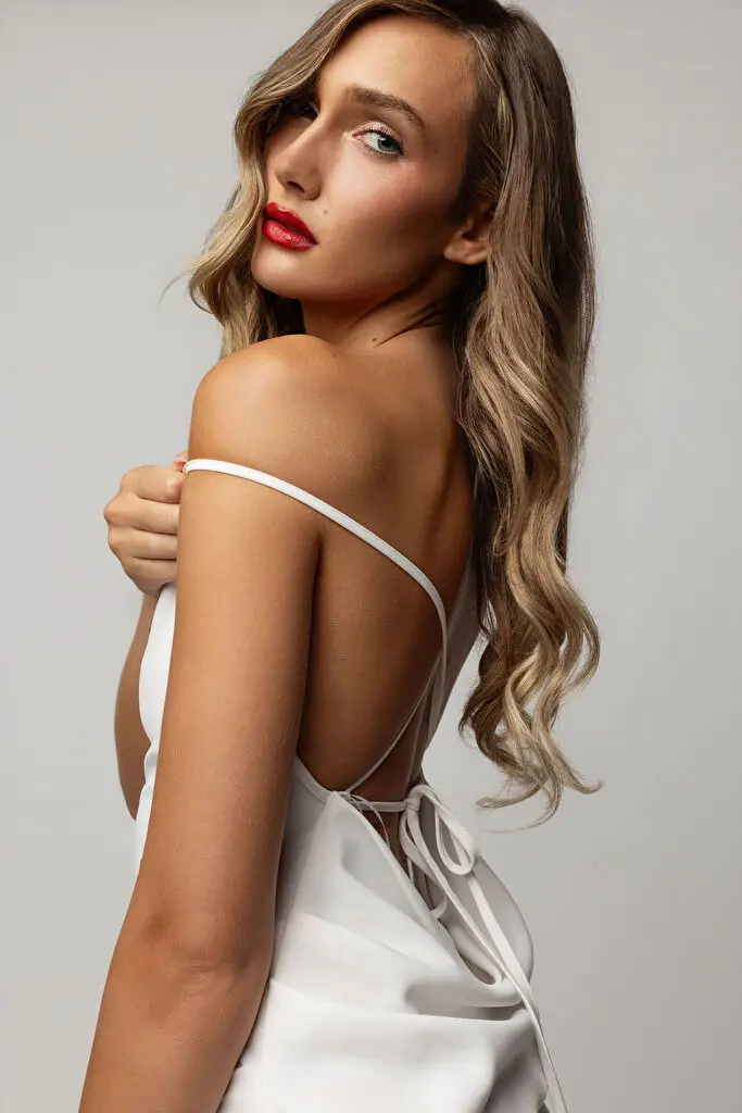 Hannah Caddo stands with her back to the camera, looking over her shoulder into the lens, one hand easing the thin strap of a white open-back satin dress off her shoulder, long blonde balayage curls to her waist, red lipstick, soft grey studio light.