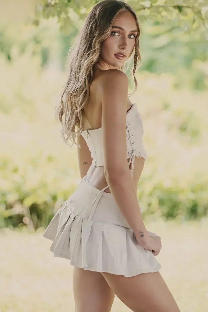 Hannah Caddo stands turned away, looking back over her shoulder, her long wavy dark blonde hair falling past her shoulder blades, in a cream strapless corset with rear lacing and a short pleated skirt whose hem she holds, amid blurred sunlit greenery.